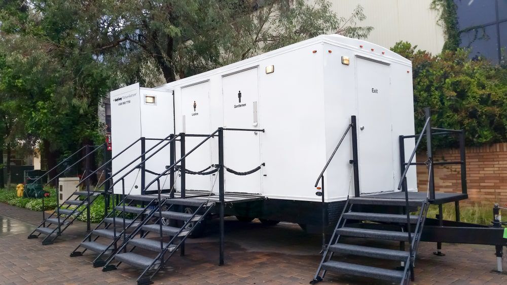 White three-stall VIP restroom trailer with stair access, set against a background of trees.