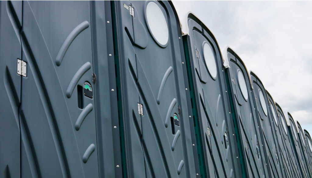 Row of gray portable toilets lined up outdoors on a cloudy day.