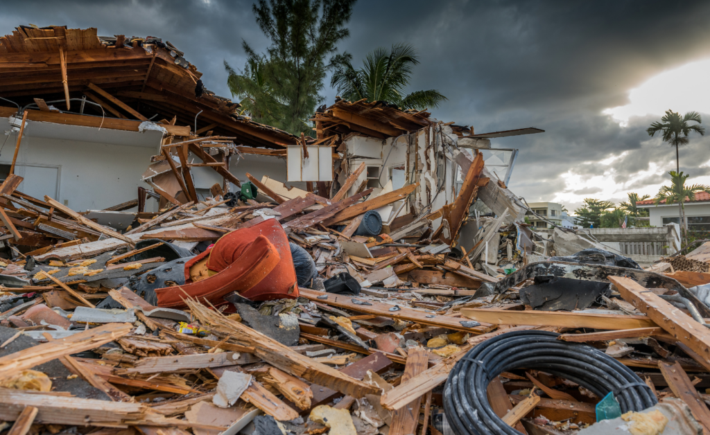 Debris from a destroyed house after a hurricane, with scattered wood, furniture, and other materials amidst dark, stormy skies and palm trees in the background.