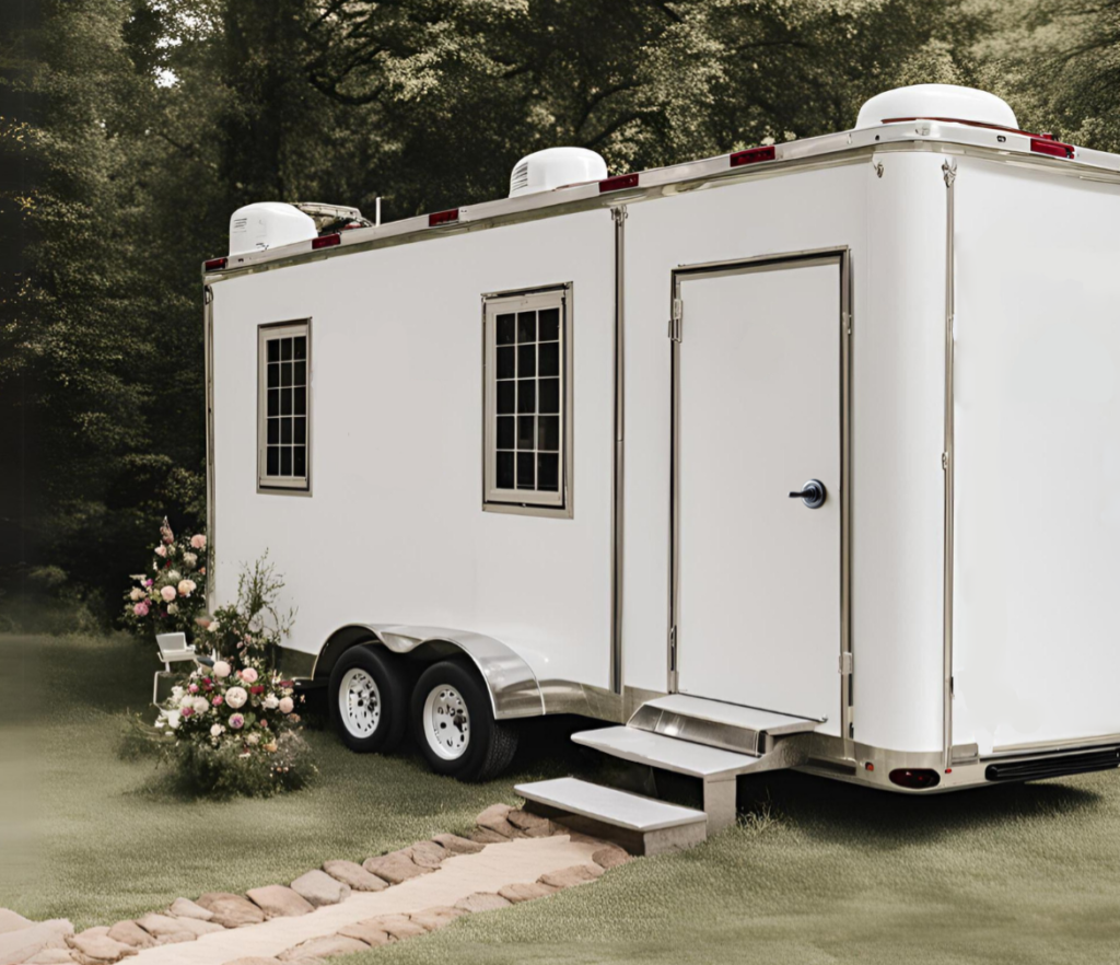 Elegant white restroom trailer set up in a lush outdoor wedding venue with flowers and greenery surrounding the area.